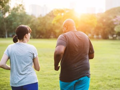 Two people, a man and woman, wear exercise gear outdoors in a green park and walk away from the camera.